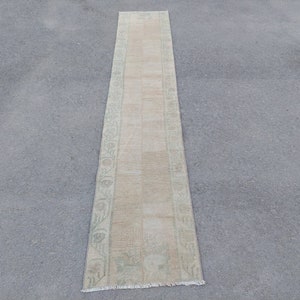May include: A long, narrow, beige runner rug with a green floral pattern along the edges. The rug has a fringed edge.