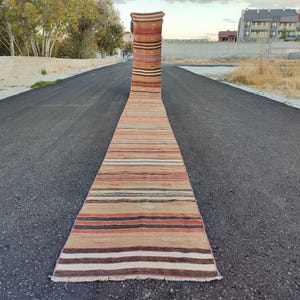 May include: A long, rectangular runner rug with a striped pattern in shades of beige, brown, orange, and white. The rug is placed on a paved road, showcasing its length and design. The rug's fringe is visible at the end.