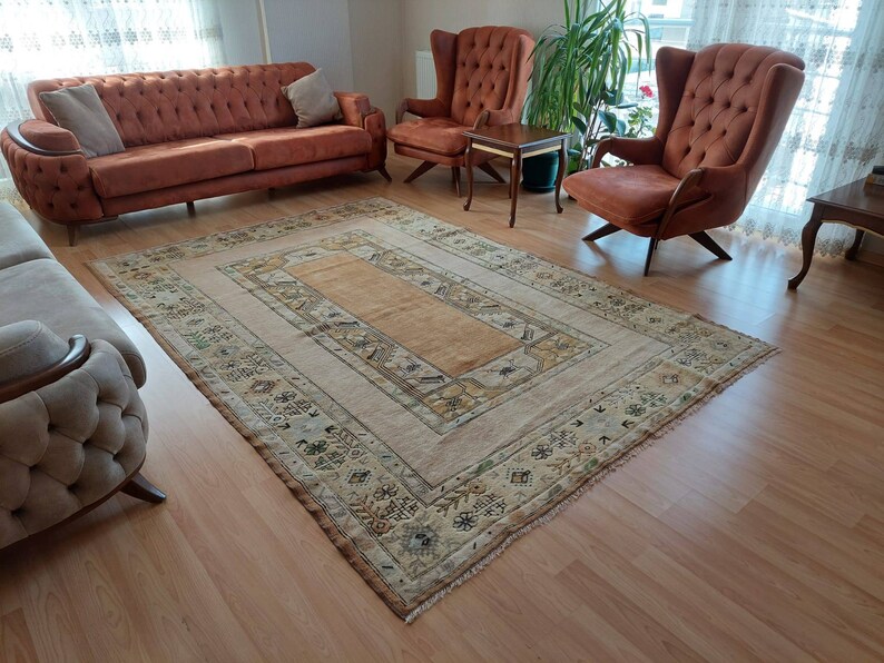 Puede incluir: Una alfombra beige y marr&oacute;n con estampado y un centro marr&oacute;n claro, colocada sobre un suelo de madera dura en un sal&oacute;n. Dos sillones marrones y un sof&aacute; marr&oacute;n est&aacute;n dispuestos alrededor de la alfombra.