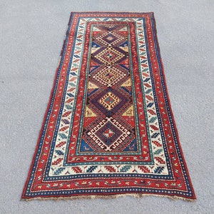 Puede incluir: Una alfombra tradicional de color rojo, azul y crema con un patrón geométrico. La alfombra tiene un borde con flecos y está hecha de lana.