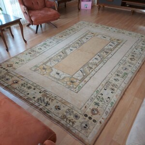 Puede incluir: Una alfombra de color crema con un centro beige y un borde estampado. La alfombra tiene un dise&ntilde;o geom&eacute;trico con peque&ntilde;os motivos repetidos en tonos marrones, verdes y beige.