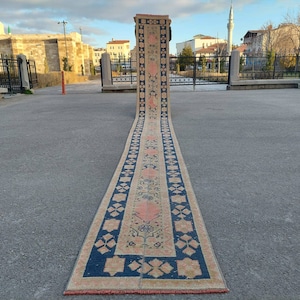 May include: A long, narrow rug with a traditional Turkish design. The rug is beige with blue and pink accents. It has a floral pattern and geometric shapes.
