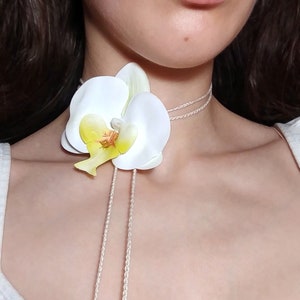 May include: A white orchid flower choker necklace with a yellow and brown center. The necklace features a delicate, braided cord that hangs down the front. The choker is a unique piece of jewelry.