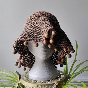 May include: A brown crocheted hat with a wide brim and wooden ball accents. The hat is displayed on a gray mannequin head. The hat's brim is adorned with evenly spaced wooden balls. The hat is a unique accessory.