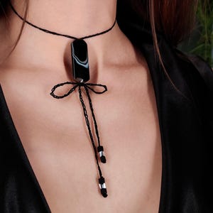 May include: A black choker necklace featuring a rectangular pendant with white and black marbling. The pendant hangs from a thin black cord, with a bow detail and dangling ends adorned with silver beads. The necklace is worn on a person.