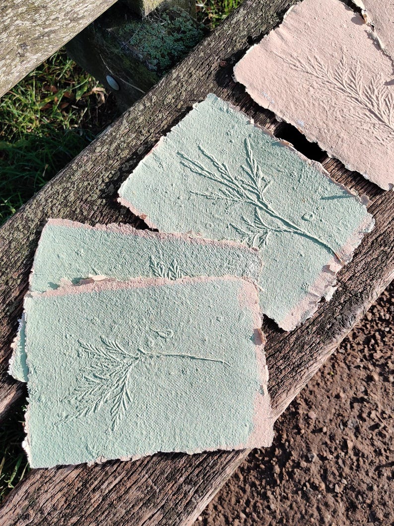 May include: Handmade paper sheets in shades of green and pink, each featuring an embossed botanical design. The paper has a textured surface and irregular edges, suggesting a natural, artisanal craft. The sheets are arranged on a weathered wooden surface.