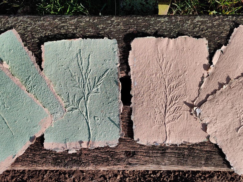 May include: Handmade paper sheets in shades of green and pink, each featuring an embossed botanical design. The edges are torn, giving a rustic, textured appearance. The paper is arranged on a weathered wooden surface.