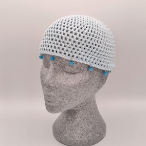 Pode incluir: Um chapéu de crochê azul claro com um design de malha. O chapéu é adornado com pequenas contas quadradas turquesas ao longo da borda inferior. O chapéu é exibido em uma cabeça de manequim cinza sobre um fundo branco.