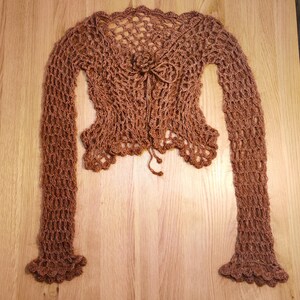 May include: A brown, open-weave crochet bolero with long sleeves. The bolero features a scalloped edge, a small flower detail at the neckline, and ties at the center. The sleeves have a flared, ruffled cuff. The garment is displayed on a wooden surface.