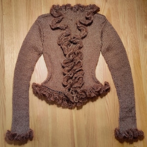 May include: A brown knitted cardigan with a ruffled neckline and cuffs. The cardigan is made of a soft, lightweight yarn and has a relaxed fit.