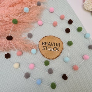 Könnte beinhalten: Eine weiße Schnur mit bunten Bommeln in Rosa, Blau, Grün und Braun. Die Schnur hängt an einem hellblauen strukturierten Hintergrund. Ein Holzschild mit dem Text "BRAVUR STOCK" befindet sich in der Mitte des Bildes.