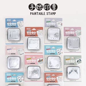 May include: A set of ten white square paint-able stamps with black outlines. The stamps feature various designs, including a cat, a calendar, a notebook, a shopping bag, a mountain range, and a blank frame. Each stamp is packaged in a clear plastic case with a colorful background and the text "手账印章" (which translates to "hand-account stamp") and "PAINTABLE STAMP".