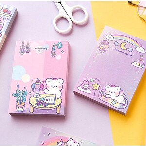 Puede incluir: Un conjunto de blocs de notas adhesivas de color rosa pastel y morado con osos de dibujos animados adorables con el texto "Hongmeimeibear". Las notas est&aacute;n decoradas con estrellas, nubes y arco&iacute;ris.