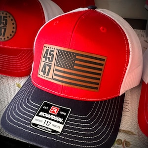 May include: Red, white, and blue trucker hat with a brown leather patch featuring the American flag and the numbers 45 and 47. The hat is adjustable and has a Richardson logo.