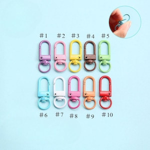 May include: A collection of ten colorful metal swivel clasps in various shades, including purple, pink, yellow, brown, and green. Each clasp has a ring and a swivel hook. The clasps are numbered from #1 to #10.