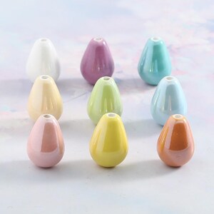 Peut inclure: Collection de neuf perles en céramique de différentes couleurs pastel, dont blanc, rose, jaune, vert et bleu. Chaque perle a une forme de goutte d'eau avec un petit trou en haut, pour l'enfilage. Les perles sont disposées sur un fond clair.