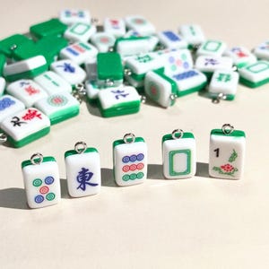 May include: A collection of small, rectangular mahjong tile charms made of white and green plastic. Each tile features a silver metal jump ring at the top for attachment. The tiles display various traditional mahjong symbols, characters, and numbers in blue, red, and green ink, with many tiles scattered in the background.
