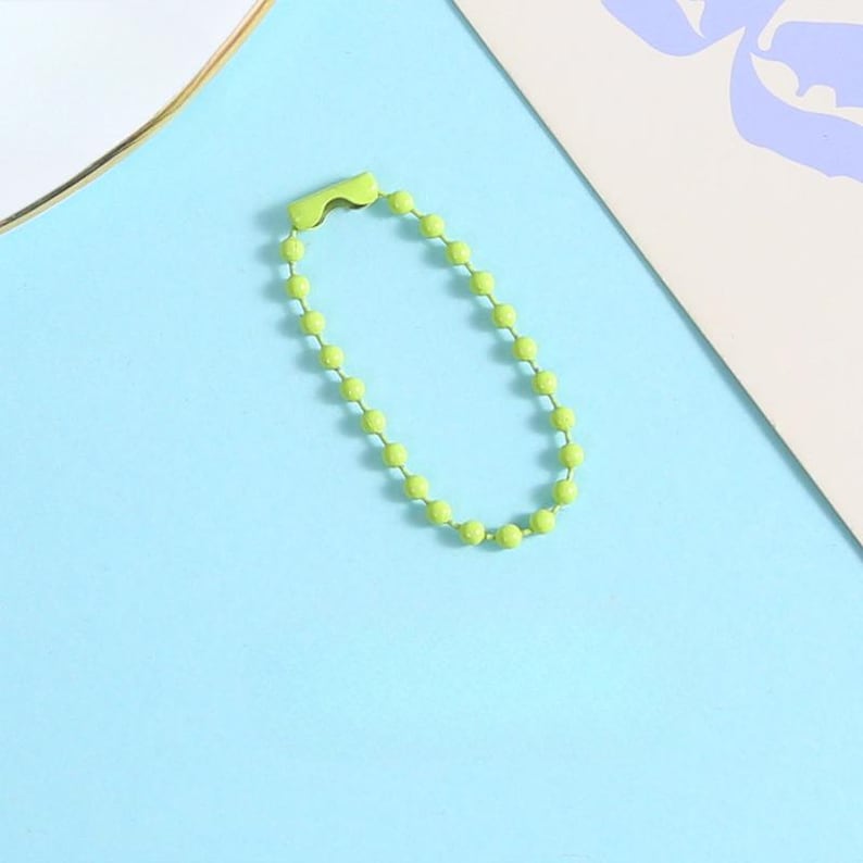 May include: A lime green ball chain with a matching clasp. The chain is composed of small, evenly spaced spheres. The background is a light blue surface with a white and beige object in the upper left corner.