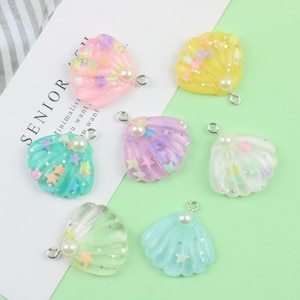 May include: A collection of colorful seashell charms in various pastel hues, including pink, yellow, green, and blue. Each charm features a pearl accent and star-shaped glitter. The charms have a silver-colored loop for attaching to jewelry or other crafts.