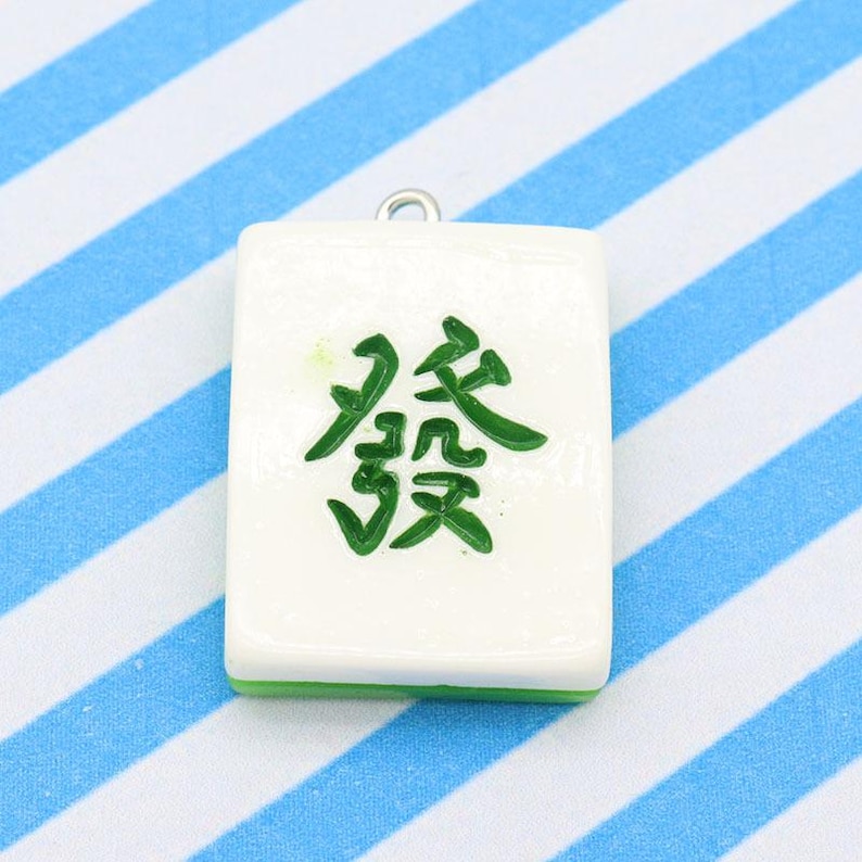 May include: A white Mahjong tile pendant with green trim and a green Chinese character. The pendant has a silver-colored loop at the top for attaching to a necklace or bracelet. The background is a blue and white striped pattern.