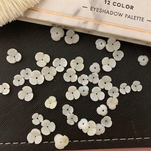 May include: A collection of white, shell-shaped flower buttons. The buttons are scattered on a black surface.