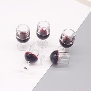 May include: Five miniature clear glass wine glasses filled with red wine. The glasses are arranged on a white and gray surface.