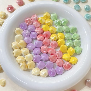 May include: A collection of pastel-colored seashell-shaped beads in a white bowl. The beads are in shades of pink, yellow, green, and purple.