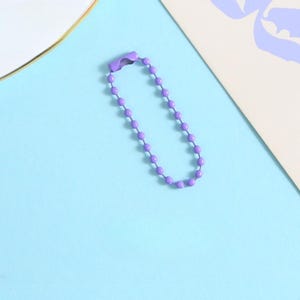 May include: A purple ball chain keychain with a clasp. The chain is made of small, evenly spaced, round beads. The background is a light blue surface with a white and purple design in the corner.
