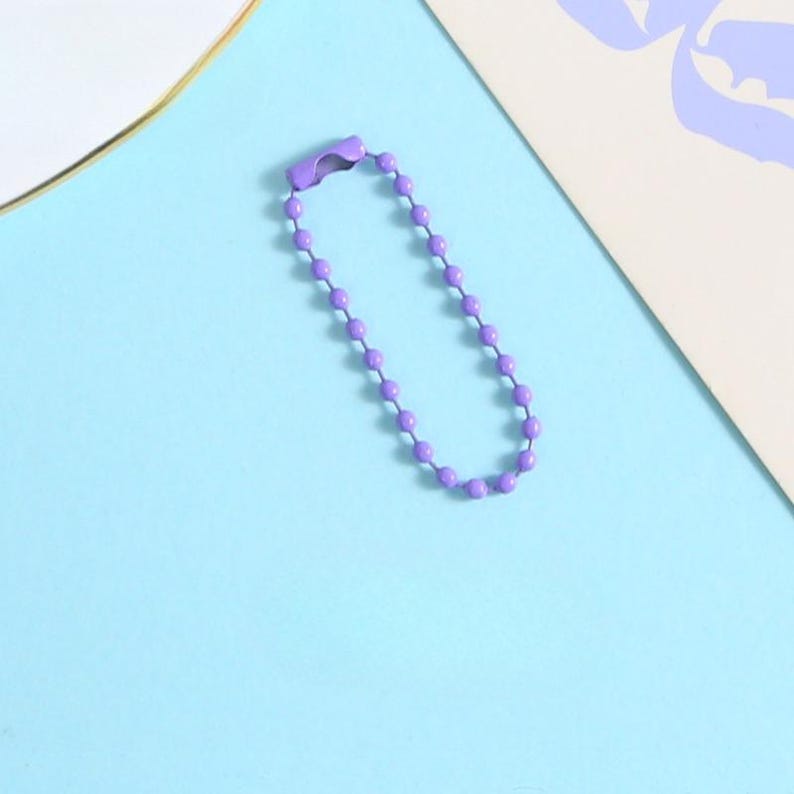May include: A purple ball chain keychain with a clasp. The chain is made of small, evenly spaced, round beads. The background is a light blue surface with a white and purple design in the corner.