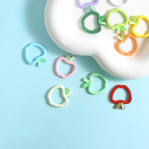 Può includere: Una collezione di ciondoli a forma di mela colorati in varie tonalità, tra cui blu, rosa, verde, giallo, arancione e rosso. Ogni ciondolo presenta un dettaglio a forma di foglia. I ciondoli sono disposti su un piatto bianco e una superficie azzurra.