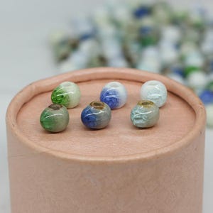 May include: Six ceramic beads in various shades of blue, green, and white. The beads are arranged in a circle on a light pink surface.
