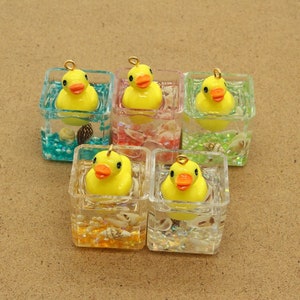 Peut inclure: Cinq breloques en forme de cube transparent contenant des canards en caoutchouc jaunes. Chaque breloque a une boucle dorée en haut et est remplie de paillettes colorées et de coquillages.