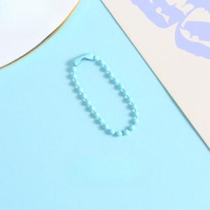 May include: A light blue ball chain keychain on a light blue background. The keychain is made of small, round beads connected together. The background also includes a white and beige paper.