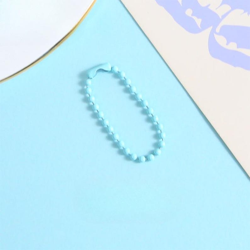 May include: A light blue ball chain keychain on a light blue background. The keychain is made of small, round beads connected together. The background also includes a white and beige paper.