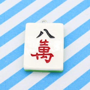 May include: A white Mahjong tile pendant with black and red Chinese characters. The tile has a green base and a silver loop for hanging. The background is a blue and white striped pattern.