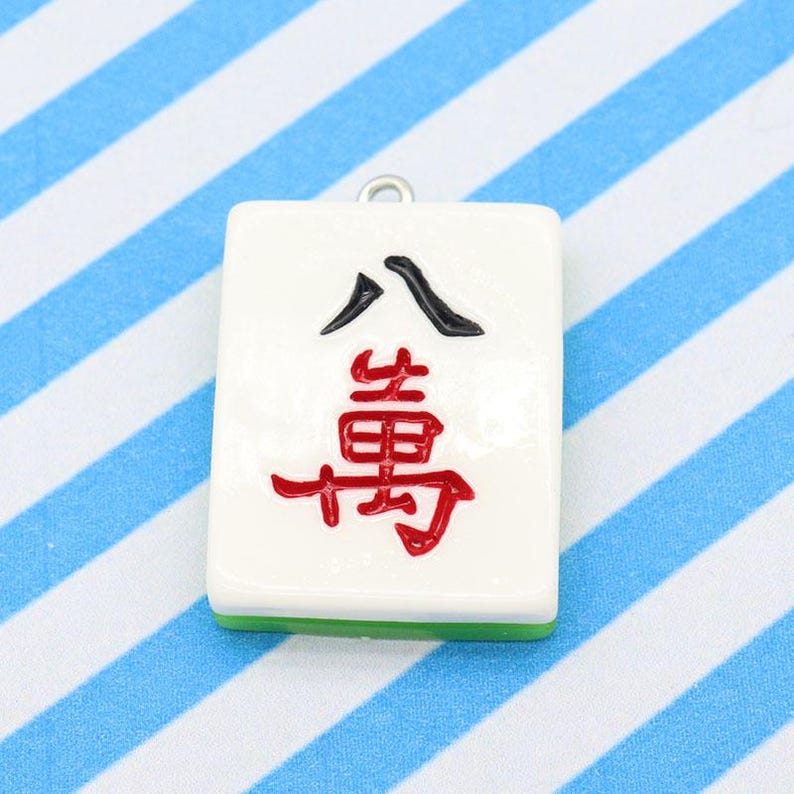 May include: A white Mahjong tile pendant with black and red Chinese characters. The tile has a green base and a silver loop for hanging. The background is a blue and white striped pattern.