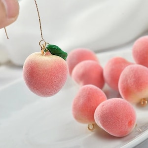 May include: Peach-shaped earrings with a green leaf charm. The earrings are made of a soft, fuzzy material and are a light pink colour. The earrings are hanging from a gold chain.