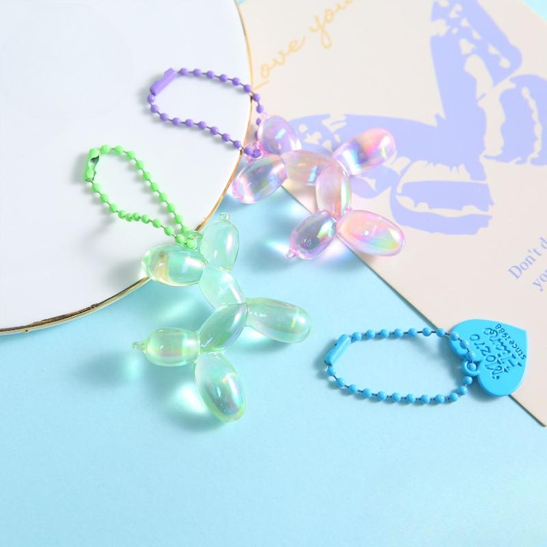 May include: Three iridescent balloon dog keychains in green, purple, and pink. Each keychain has a ball chain and a clear, dog-shaped charm. A blue heart-shaped charm with text is also visible.