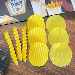 Peut inclure: Un assortiment de breloques jaunes sur le thème de la nourriture, comprenant des frites ondulées et des chips. Les breloques sont disposées sur une surface en bois, avec une boîte de ketchup et un contenant de frites en arrière-plan.