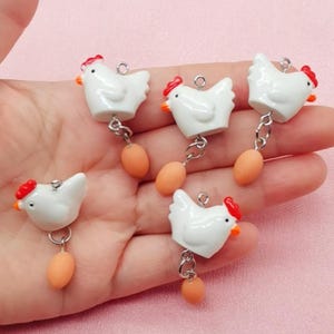 May include: A collection of white chicken charms with red combs and orange beaks, each dangling a small, peach-colored egg. The charms are connected by silver-colored chains. The overall theme is farm animals.