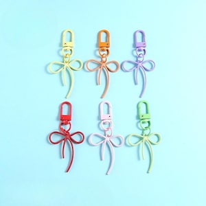 May include: Six colourful keychains with bow-shaped charms and lobster claw clasps. The keychains are in yellow, orange, purple, red, pink, and green. The background is a light blue.