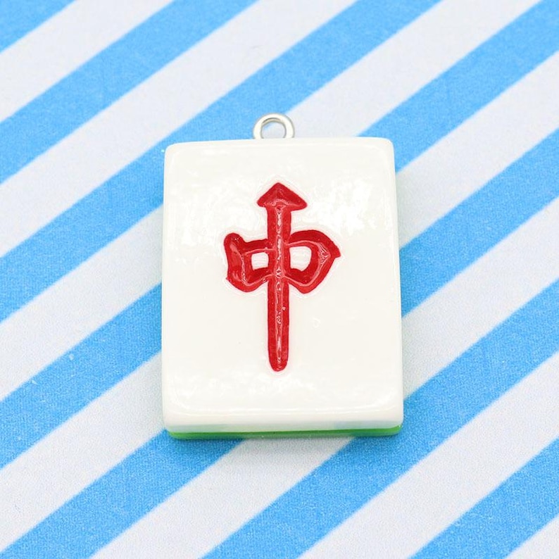 May include: A white rectangular Mahjong tile pendant with a red Chinese character. The tile has a green edge and a silver loop for hanging. The background is a blue and white striped pattern.