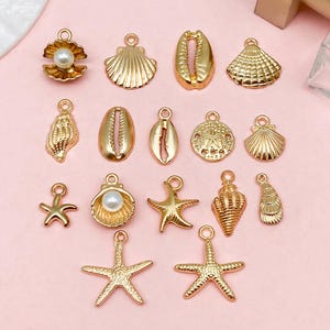 May include: A collection of gold-toned metal charms featuring various sea-themed shapes including starfish, conch shells, scallops, and cowrie shells. Two of the scallop shell charms are accented with a single white faux pearl bead.