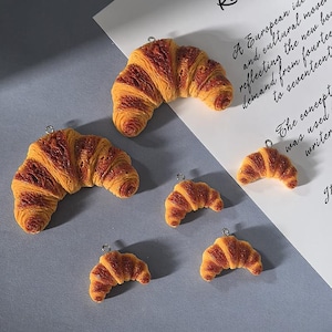 May include: Several miniature croissant charms in shades of brown and yellow are scattered across a gray surface. Each charm has a small silver loop at the top. A piece of paper with handwritten text is in the background.