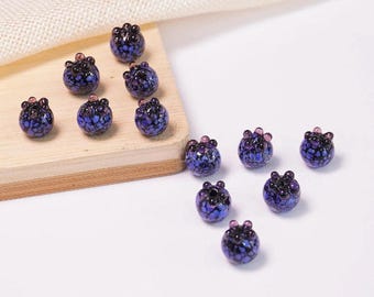 May include: A collection of glass beads shaped like blueberries. The beads are primarily dark blue with speckled patterns and small pink accents on top. Some beads are arranged on a small wooden block, while others are scattered on a white surface.