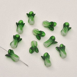May include: A collection of 12 green and white glass beads shaped like bok choy vegetables. The beads are approximately 2.5 cm in length and have a glossy finish.