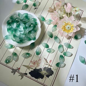 May include: A collection of translucent green leaf-shaped beads scattered on a patterned surface. A white bowl holds a pile of the beads. A floral arrangement with pink and yellow accents is also present. The image is labeled with the number "#1".