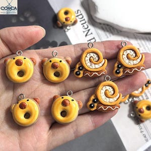 May include: A collection of yellow donut-shaped charms with brown bear ears and noses, and snail-shaped charms with white swirls. Each charm has a silver loop for attaching to jewelry or other crafts.
