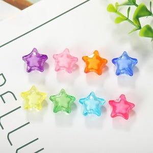 May include: Eight translucent star-shaped beads in various colors: purple, pink, orange, blue, yellow, green, light blue, and red. The beads are arranged on a white surface, with a few green leaves in the background.