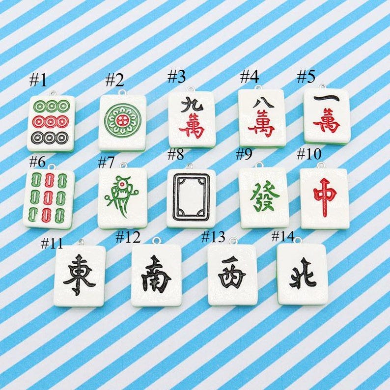 May include: A collection of fourteen white Mahjong tile pendants with black, red, green, and gold symbols and characters. Each tile has a silver loop for hanging. The background is a blue and white striped pattern.